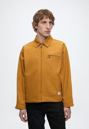 Ung mand med lysebrunt krøllet hår iført en sennepsgul lynlåstrøje med en brystlomme med lynlås og sorte bukser.