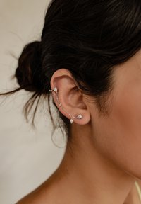 Srebrny klips na ucho z łańcuszkiem, zdobiony trzema przejrzystymi akcentami kamieni szlachetnych. Gładka tekstura i zaokrąglony kształt, umieszczone na teksturowanym tle.