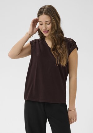 Młoda kobieta z długimi falującymi włosami, ubrana w ciemnobrązowy, bezrękawnik z koronkowymi ramionami i czarne spodnie, dotyka włosów i delikatnie się uśmiecha.