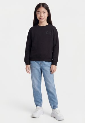 Girl with long black hair wearing a black sweatshirt with "CK" detail, light blue jeans, and white sneakers, standing against white background.