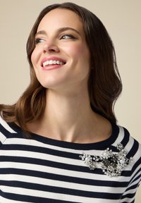 Ein navy- und weiß gestreifter Pullover mit einer dekorativen Brosche aus Perlen und Strass auf der linken Schulter. Glattes Gewebe, runder Ausschnitt.