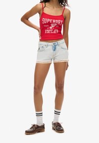 Rode tanktop met witte letters, denim shorts met lichte wassing en kanten rand, witte sokken met zwarte strepen, bruine schoenen met een robuuste zool.