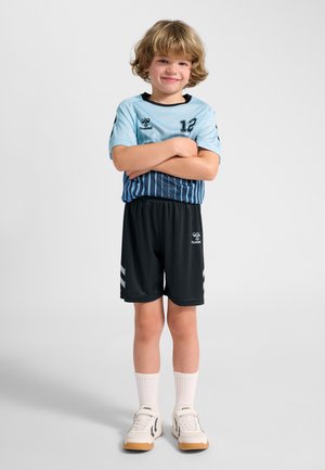 Dziecko o blond włosach stoi z założonymi ramionami, ubrane w jasnoniebieską i czarną sportową koszulkę z numerem 12, czarne spodenki, białe skarpetki i trampki.