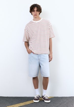 Tânăr cu păr creț purtând un tricou oversized alb cu dungi roșii, pantaloni scurți din denim albastru deschis, șosete albe și adidași închiși la culoare.