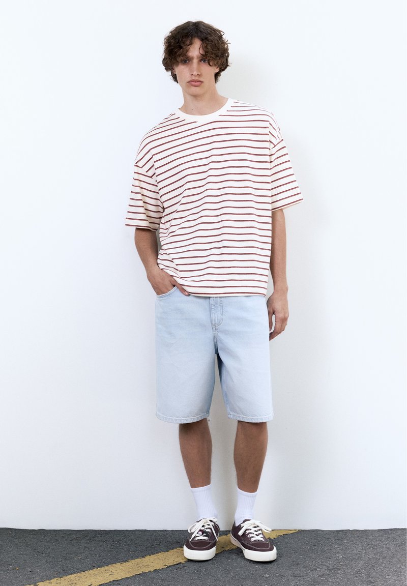 Jeune homme aux cheveux bouclés portant un t-shirt oversize blanc à rayures rouges, un short en jean bleu clair, des chaussettes blanches et des baskets foncées.