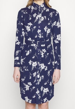 Femme portant une robe bleu marine boutonnée avec imprimé floral blanc et ceinture assortie, debout devant un fond blanc uni.