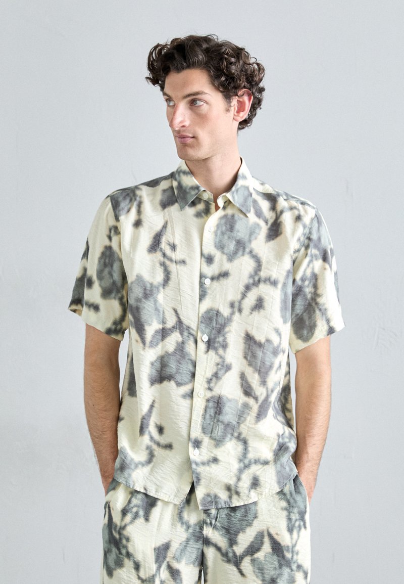 Short-sleeve, collared shirt in light beige with black and gray abstract patterns. Made of textured fabric, featuring a button front and relaxed fit.
