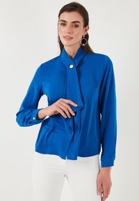 Bluză de culoare albastru cobalt cu guler înalt, detaliu cu fundă drapată, mâneci lungi și accente din butoane argintii; material fin și croială ajustată.