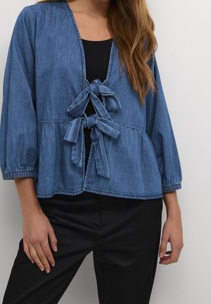 Vrouw draagt een blauwe spijkerjack met twee strikken aan de voorkant, over een zwarte top en zwarte broek, staand tegen een witte muur.