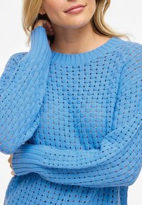 Femme portant un pull en maille bleu clair avec un motif texturé et perforé, un bras croisé et l'autre main touchant son cou.