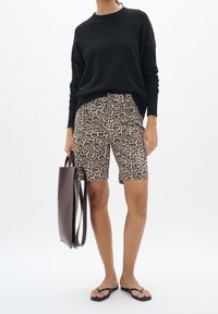 Femme portant une chemise noire à manches longues, un short imprimé léopard, des tongs noires, tenant un sac fourre-tout en cuir brun foncé à côté d'elle.