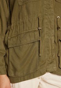 Lekka oliwkowo-zielona kurtka wykonana z trwałego materiału, z dwoma przednimi kieszeniami, paskiem ze ściągaczem w talii oraz metalowymi elementami do regulacji.