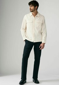 Light beige button-up shirt with two front pockets, textured pattern, paired with black trousers and black dress shoes.