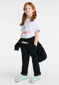 Weißes T-Shirt mit buntem Fisch-Motiv, dunkle Hose und grüne kariert gemusterte Slipper. Schwarzer Pullover über den Schultern.