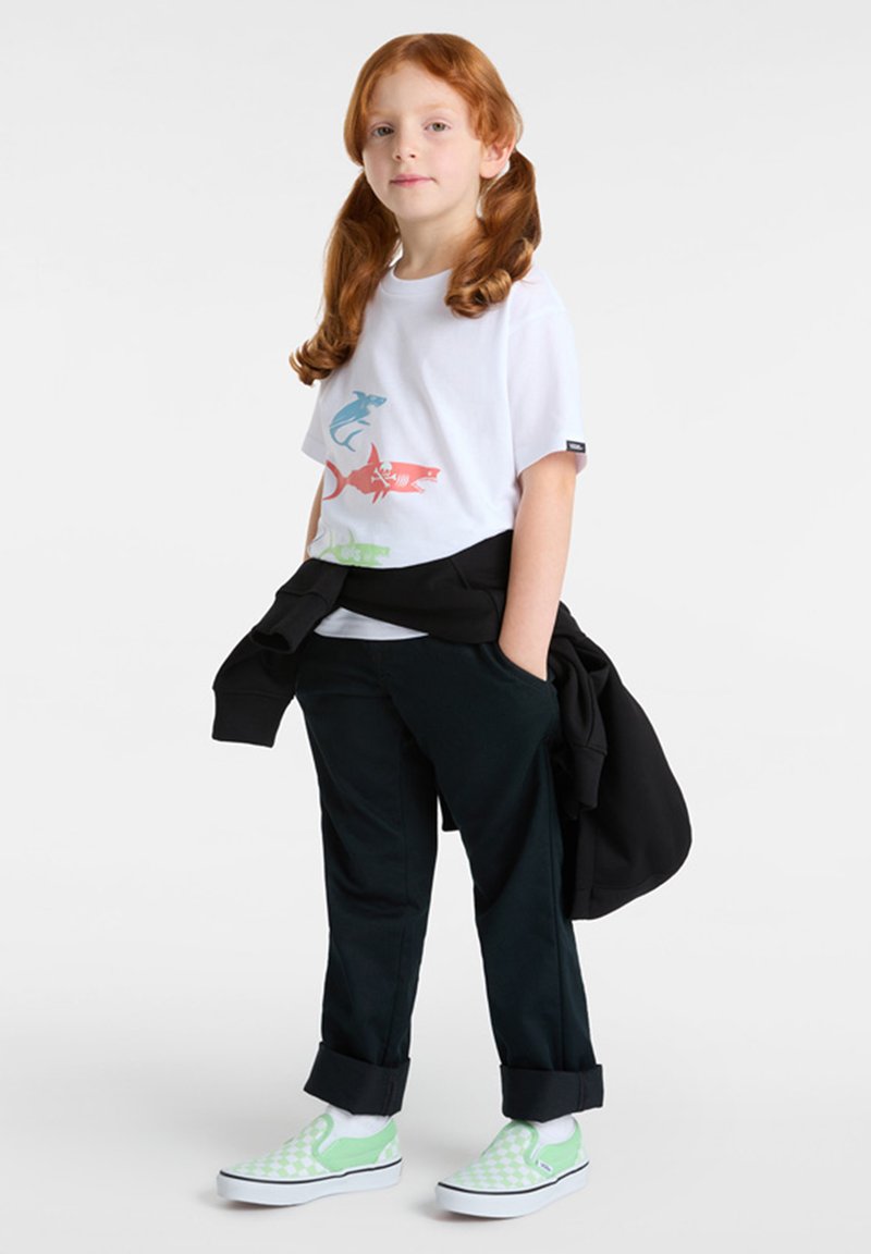 Weißes T-Shirt mit buntem Fisch-Motiv, dunkle Hose und grüne kariert gemusterte Slipper. Schwarzer Pullover über den Schultern.