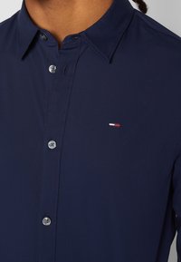 Mörkblå skjorta med knappar i slät väv. Har en klassisk krage och en liten broderad logotyp i röd, vit och blå på bröstet.