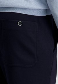 Close-up van een donkerblauwe broekzak met een zwart knoopdetail, gedragen met een lichtblauwe geribbelde trui.