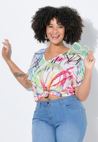V-hals, top med korte ærmer i et farverigt, abstrakt mønster. Stoffet er let med en knude foran. Modellen holder grønne solbriller og har blå jeans på.