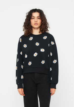 Kvinde med krøllet hår iført en sort sweatshirt med hvide margueritmønstre og sorte bukser, stående foran en ensartet lys baggrund.