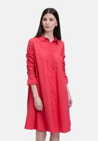 Robe chemise en coton rouge avec un devant à boutons, des manches longues avec des détails froncés et une jupe évasée. Texture lisse et design ludique.