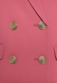 Manteau rose croisé avec quatre grands boutons ronds beige et deux poches à rabat à l'avant.