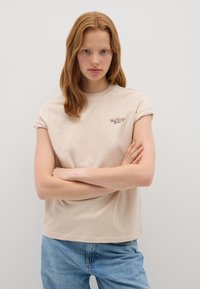 T-shirt en coton beige à manches courtes avec des manches retroussées et "SUNDAY" imprimé en marron sur la poitrine. Porté avec un pantalon en jean denim bleu clair.