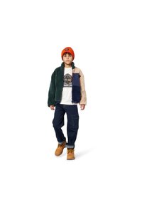 Veste en polaire multicolore avec des panneaux bleus, verts et beiges, t-shirt blanc avec logo, jeans foncés, et bonnet en tricot orange. Bottes marron.