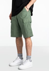 Pantalones cortos cargo en algodón verde oliva, con bolsillos laterales y un corte holgado. Combinados con zapatillas blancas y calcetines grises. Diseño simple y práctico.