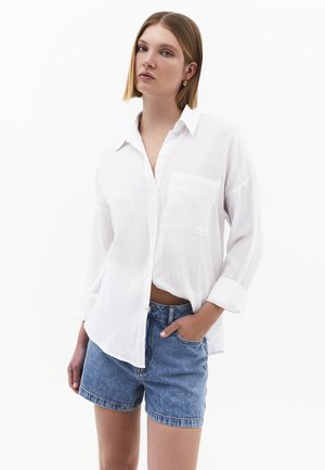 Nainen, jolla on suorat olkapäille ulottuvat hiukset, jolla on yllään löysä valkoinen nappipaita käärittyine hihoineen ja farkkushortsit, seisoo yhdellä kädellä taskussa.