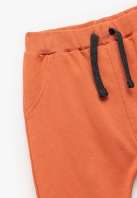 Pomarańczowe spodnie dresowe z żebrowanym paskiem w talii, czarnym sznurkiem i boczną kieszenią. Wykonane z miękkiego materiału o gładkiej fakturze.