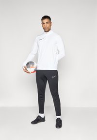 Weißes, mit Reißverschluss versehenes Sportoberteil mit schwarzem Nike-Logo, kombiniert mit schwarzen, schmal zulaufenden Hosen mit Logo, während ein schwarz-weißer Fußball gehalten wird.