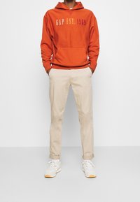 Osoba v oranžové mikině GAP s textem "EST. 1969", béžových zkrácených kalhotách a bílých teniskách stojí se spuštěnýma rukama v kapsách.