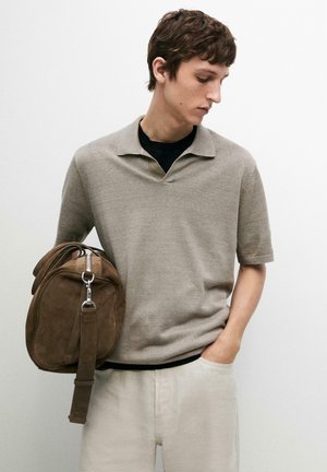 Giovane uomo con maglione beige a maniche corte e pantaloni chiari, che tiene in mano una borsa da viaggio in suede marrone, guardando verso il basso di lato su sfondo bianco.