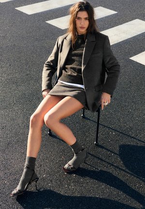 Femme en blazer gris, jupe assortie, pull et talons hauts avec chaussettes, assise sur une chaise sur l'asphalte près d'un passage piéton.