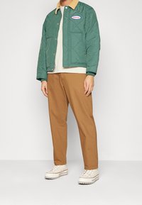 Vadderad grön jacka med en beige manchesterkrage, logotyp patch, bärs över en krämfärgad skjorta, i kombination med bruna byxor och vita sneakers.