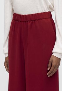 Gros plan d'une personne portant un pantalon rouge ample taille haute avec une taille élastique et un haut blanc à manches longues avec des poignets côtelés.