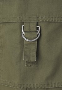 Tămbălău de culoare verde măsliniu cu o suprafață texturată, având un inel D din metal și o bandă pentru fixare, împreună cu detalii de cusătură vizibile.