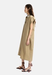 Khaki oversize kjole med korte ærmer, der har en dekorativ krave. Båret med brune sandaler pyntet med røde kvaster.