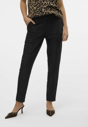 Kobieta ubrana w czarne spodnie cargo z kieszeniami na klapę, top w panterkę oraz brązowe szpilki z ozdobnymi oczkami i kokardkami.