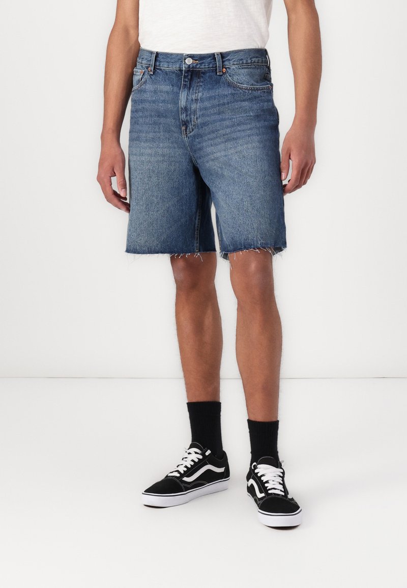 Denim shorts i en mellemblå vask, med rå kant, klassisk fem-lomme design og slidt tekstur. Matches med sorte sneakers og sokker.