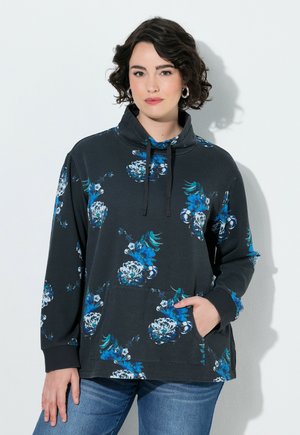 Kvinde med kort krøllet hår iført en mørk blomstret sweatshirt og blå jeans, står med den ene hånd i lommen.