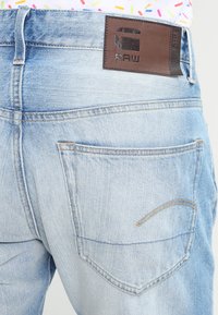 Jeans in denim azzurro chiaro con una texture scartavetrata. Presentano un'etichetta in pelle marrone sulla cintura con il marchio "RAW" e due tasche posteriori.
