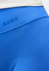 Κοντινή λήψη μπλε ριμπ ύφανσης κολάν με καμπυλωτή ραφή και ανάγλυφο κείμενο "BORG" κοντά στη μέση κάτω από λευκό τοπ.