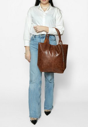 Vrouw met witte blouse en blauwe spijkerbroek houdt een grote bruine leren boodschappentas met kettingkwastdetail vast.