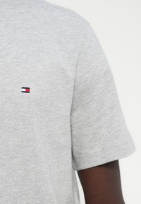 Ανοιχτό γκρι κοντομάνικο μπλουζάκι με μικρό ορθογώνιο λογότυπο σε ναυτικό μπλε, λευκό και κόκκινο στο πάνω μέρος του στήθους.