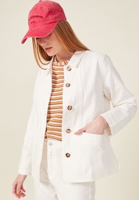 Giacca di jeans bianca con una vestibilità strutturata, caratterizzata da grandi bottoni e una tasca. Indossata su un top a righe arancione e bianco e pantaloni bianchi.