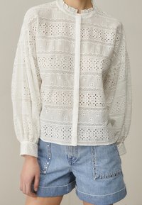 Blusa bianca ricamata con motivi in occhio di pernice, colletto ricamato e chiusura a bottoni, abbinata a pantaloni corti in denim decorati con borchie.