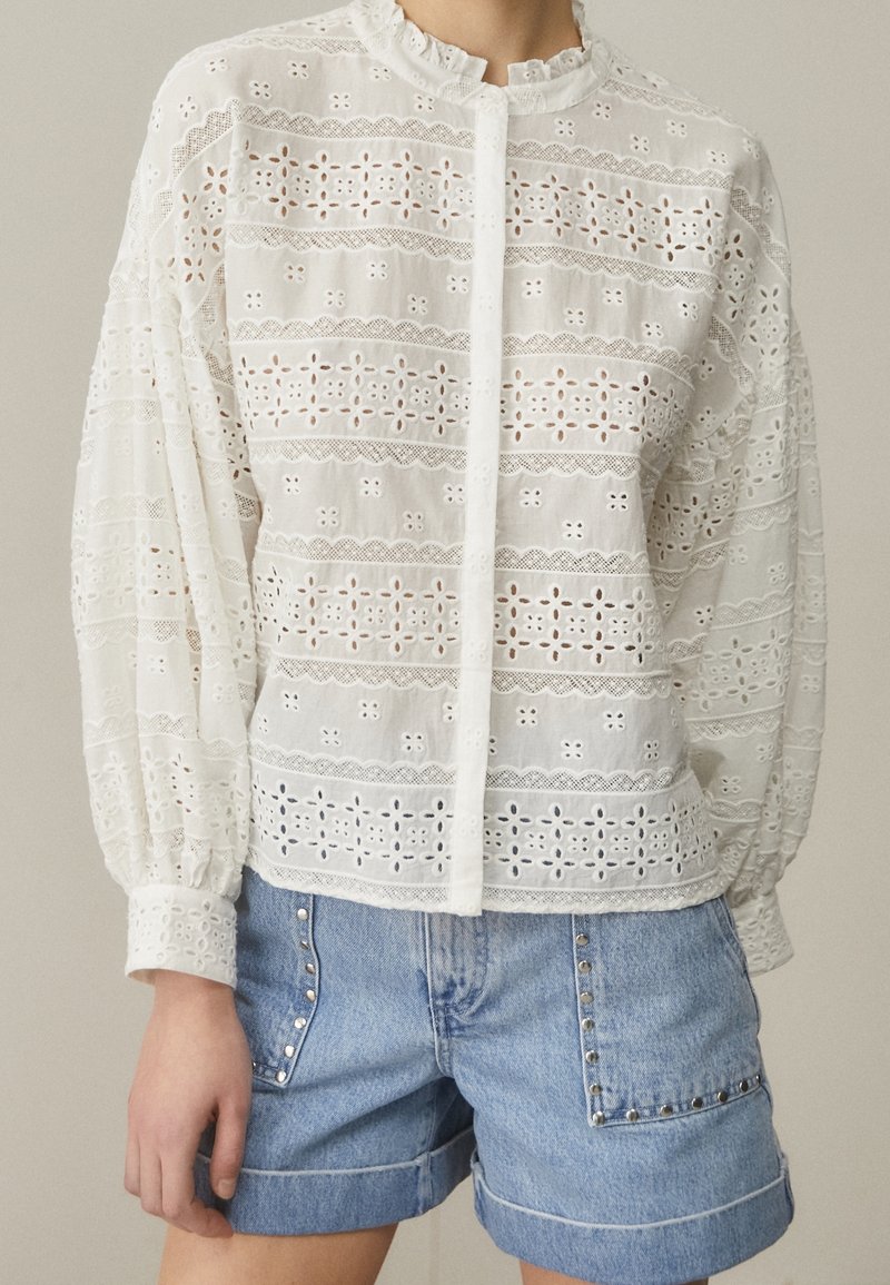Blusa bianca ricamata con motivi in occhio di pernice, colletto ricamato e chiusura a bottoni, abbinata a pantaloni corti in denim decorati con borchie.