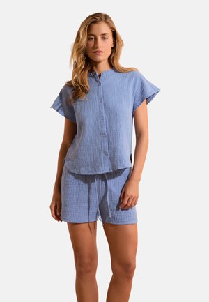 Donna che indossa un top azzurro chiaro a maniche corte con bottoni e pantaloncini coordinati con coulisse, in piedi su uno sfondo bianco semplice.