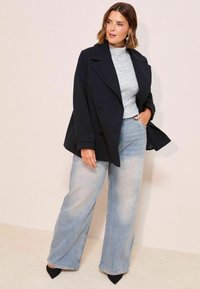Manteau peacoat noir doublé, à larges revers, porté sur un pull gris côtelé. Jean bleu clair à jambes larges et escarpins noirs à bout pointu.